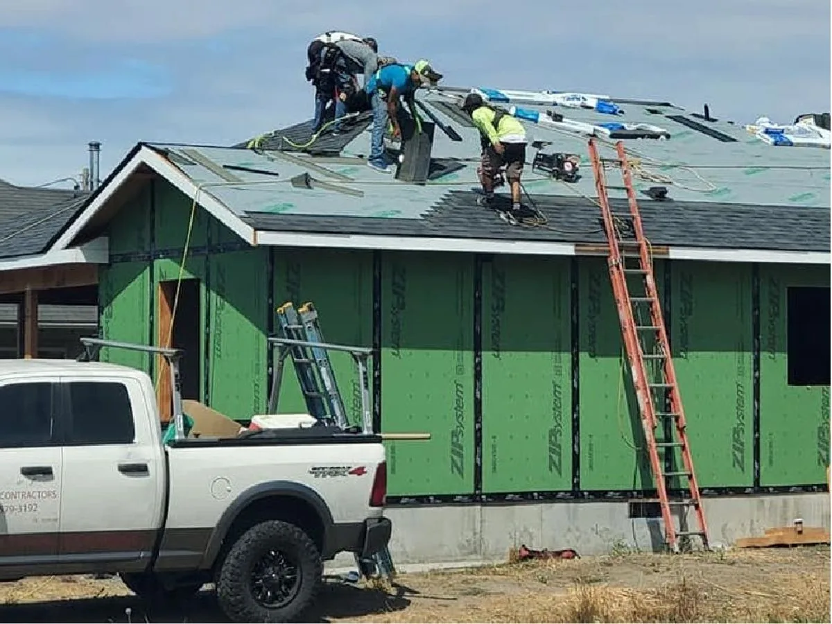 Construction workers installing a roof on a house, with tools and a truck nearby.